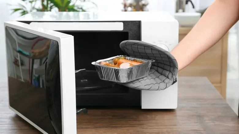 A hand wearing a gray quilted oven mitt is pulling a disposable aluminum foil tray filled with cooked food out of an open white microwave on a kitchen countertop.