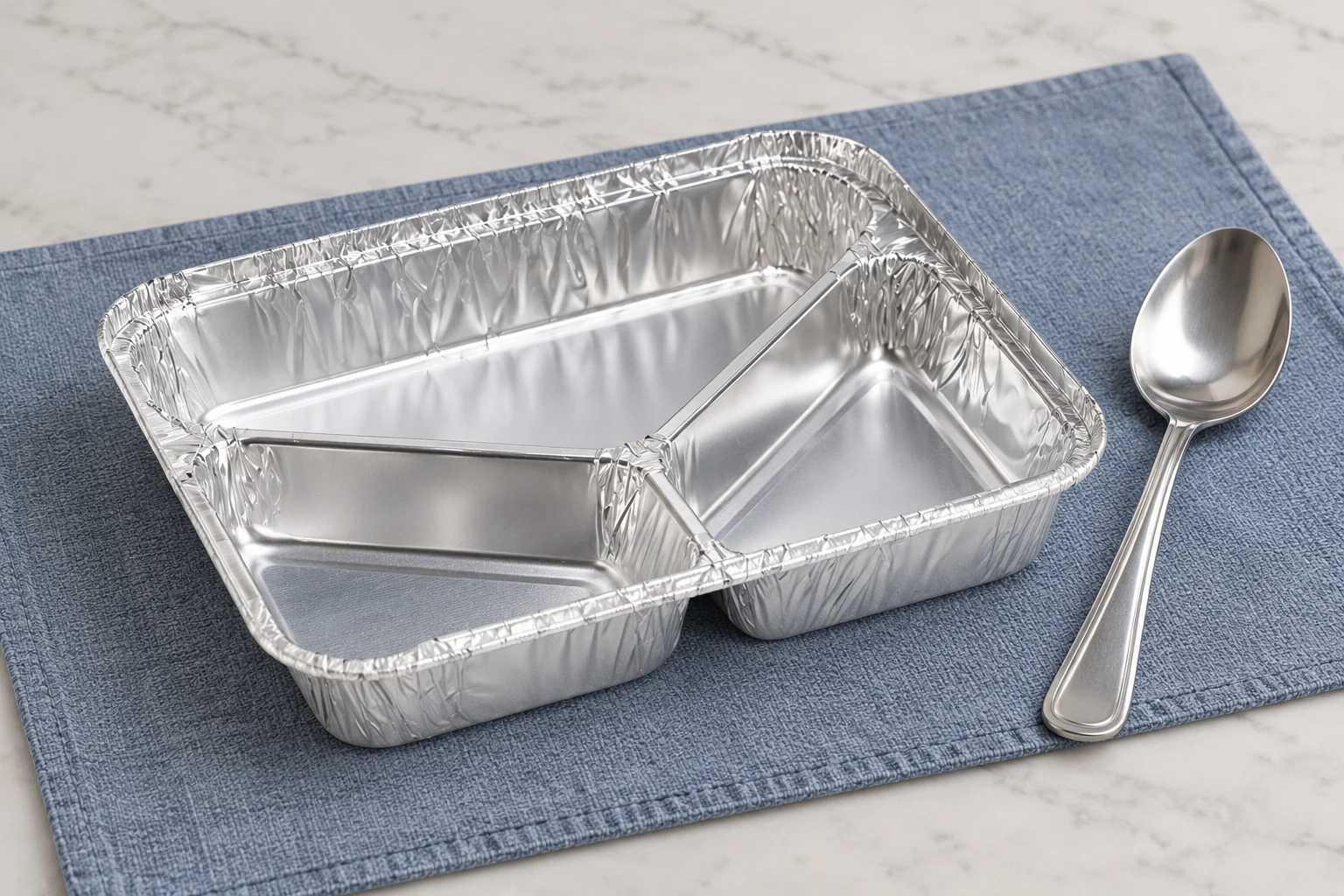 Empty three-compartment foil tray with a separate spoon on a blue-grey mat