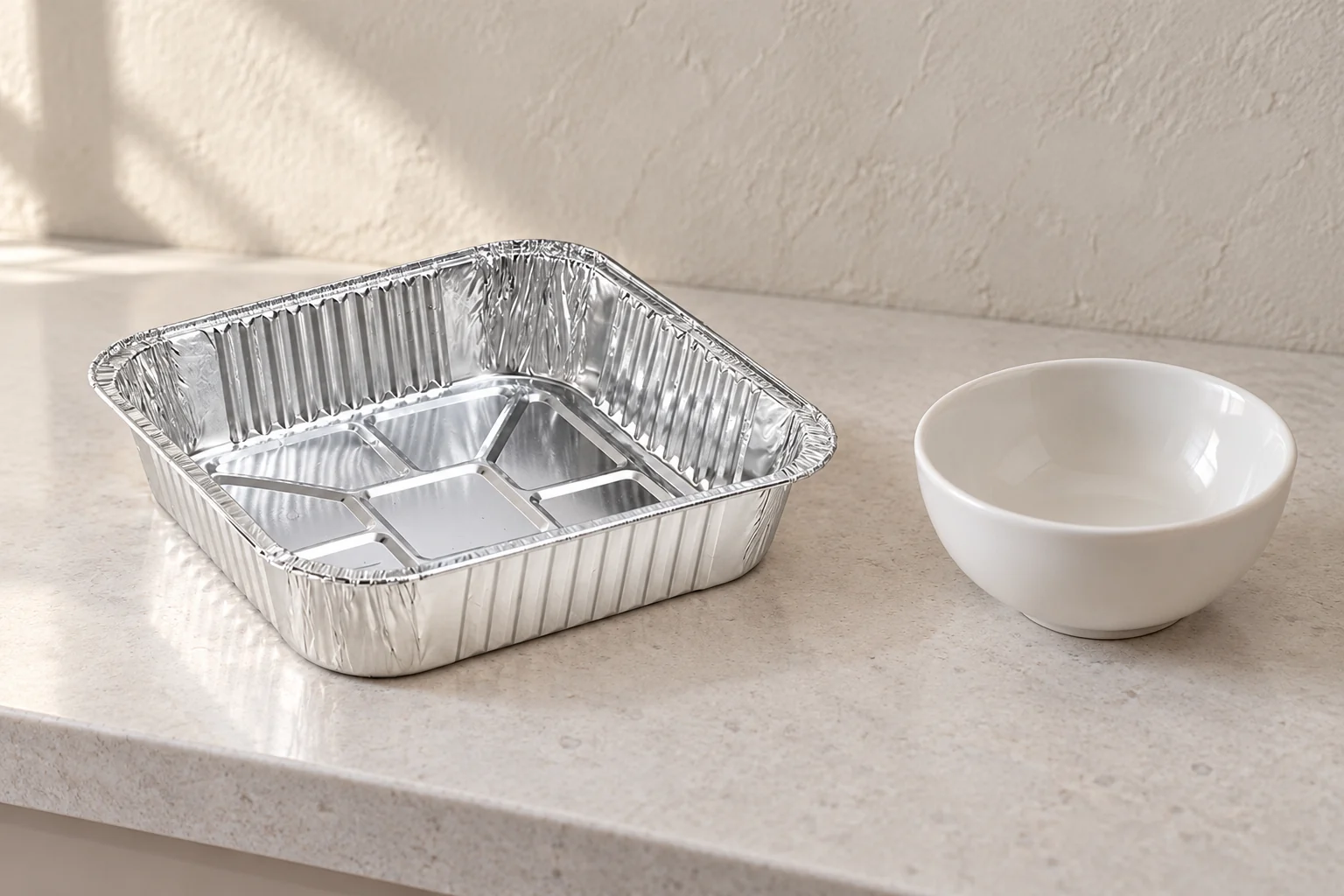 Empty square foil tray and separate empty ceramic bowl on a pale counter